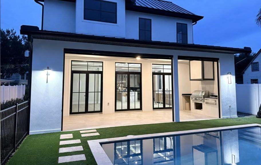 Full rear view of a two-story residence with illuminated black-framed doors beside a pool